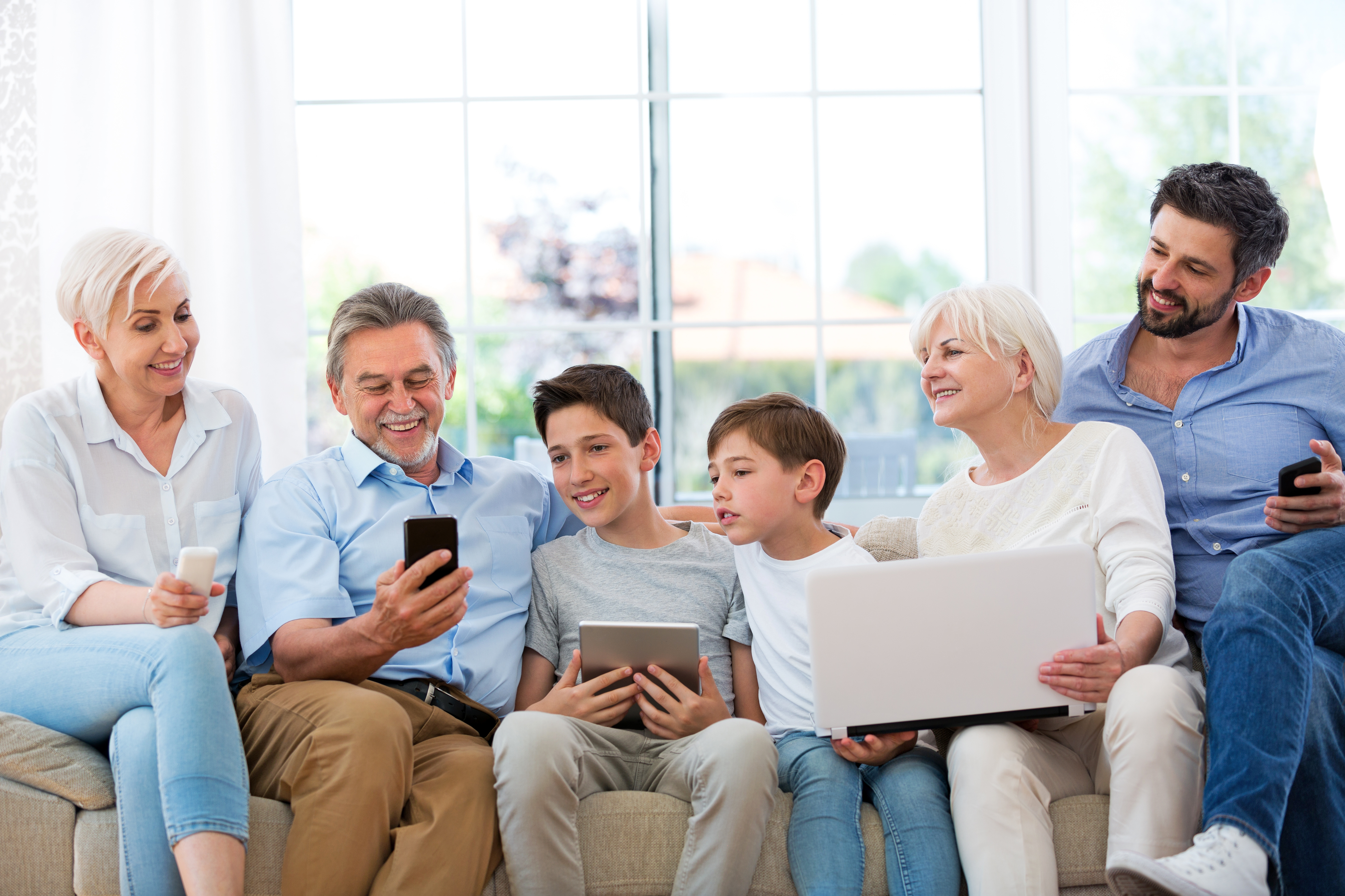 Auf dem Bild sind nebeneinander auf einem Sofa sitzende Personen verschiedenen Alters mit unterschiedlichen digitalen Endgeräten zu sehen.